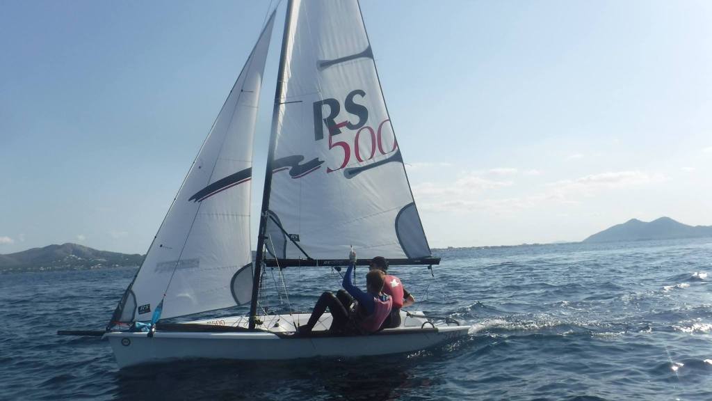 Eine weiße Segeljolle vom Typ RS500 mit zwei Personen in Neoprenanzügen und Schwimmwesten segelt auf dem Meer vor Port de Pollença auf Mallorca. Im Hintergrund Berge an der Küste und blauer Himmel.