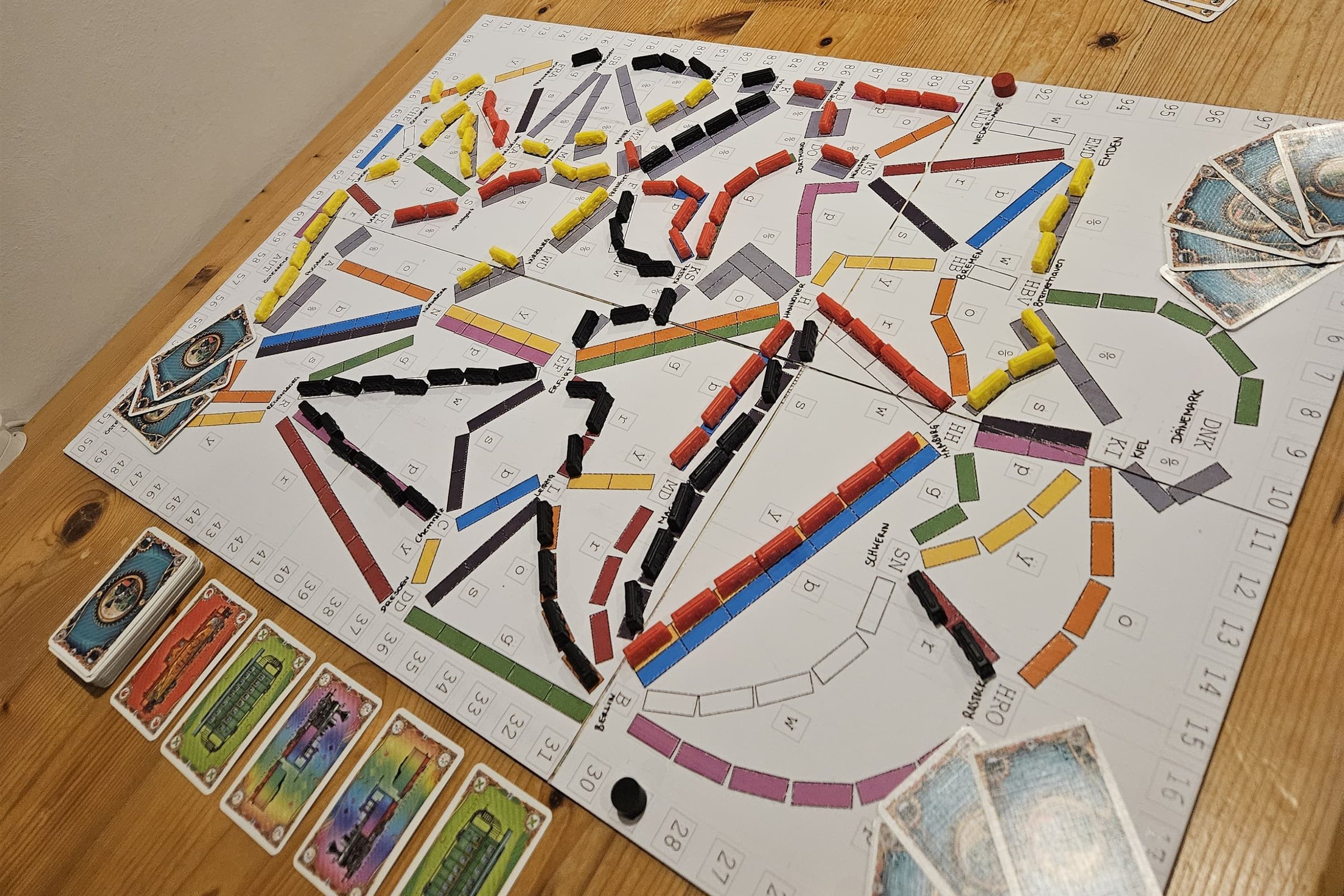Ein selbst angepasster Spielplan von Zug um Zug Deutschland auf einem Holztisch. Auf dem weißen Plan verlaufen farbige Strecken zwischen beschrifteten Städten wie Berlin, Hamburg und Kiel, darauf viele rote, gelbe und schwarze Zugwaggons. Am Rand liegen bunte Spielkarten.